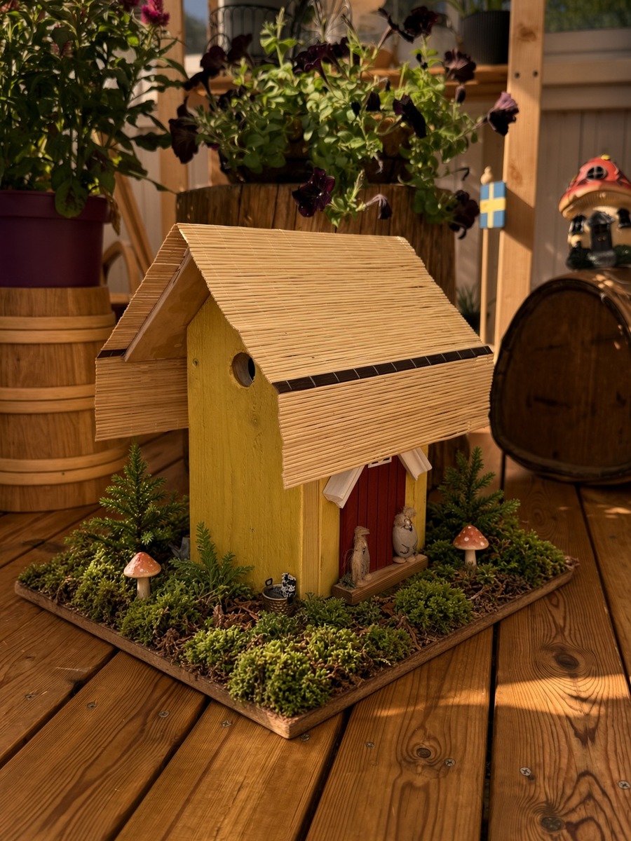 Handmade yellow wooden birdhouse with a pale corrugated roof and a small red door, standing among moss and plants
